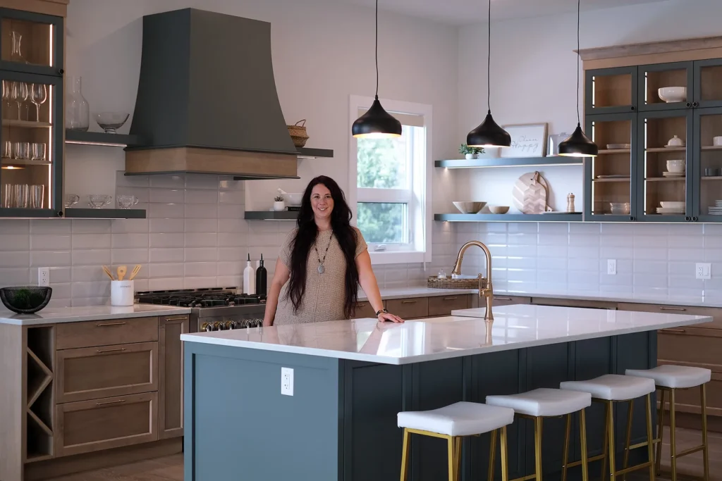 Senior Design Consultant Allison Braun standing in a modern transitional kitchen featuring a dark teal island, light wood cabinetry, white subway tile backsplash, and elegant gold hardware.