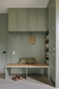 A custom sage green mudroom featuring built-in cabinetry, a wood-topped bench, and open shelving. The high-value design includes overhead storage lockers, minimalist black hooks, and integrated side cubbies with wicker baskets for organization.
