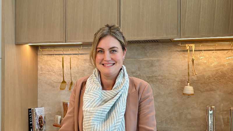 A professional portrait of Westwood Cabinetry Senior Designer Kati Knorr smiling in a modern kitchen, featuring sleek light wood upper cabinets, a stone backsplash, and gold kitchen accessories.