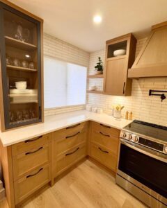 A modern custom kitchen featuring light wood cabinetry, white quartz countertops, and a white subway tile backsplash. The design includes a mix of floating wood shelves, a large glass-front display cabinet, and a custom wood range hood by Westwood Cabinetry.