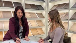 A Westwood Cabinetry designer and client in a showroom reviewing kitchen blueprints at a marble desk, with a backdrop of various wood cabinet door samples on display shelves.