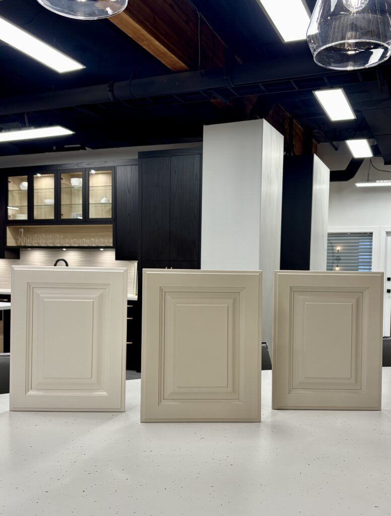 Three traditional style cabinet door samples in soft warm white and grey finishes sitting on a white speckled countertop in a modern showroom.
