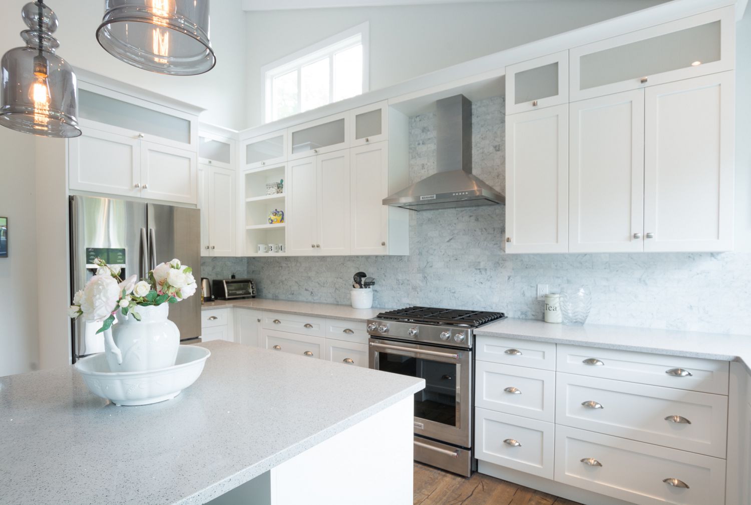 A bright minimalist kitchen designed by a kitchen design expert featuring white cabinetry with glass inserts, marble tile backsplash, and elegant pendant lighting.