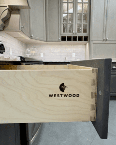 Close up view of the durable dovetail joinery on a light wood drawer box, featuring the black Westwood logo, part of high-quality custom kitchen cabinets in Kelowna in a kitchen showroom setting with grey upper cabinets.