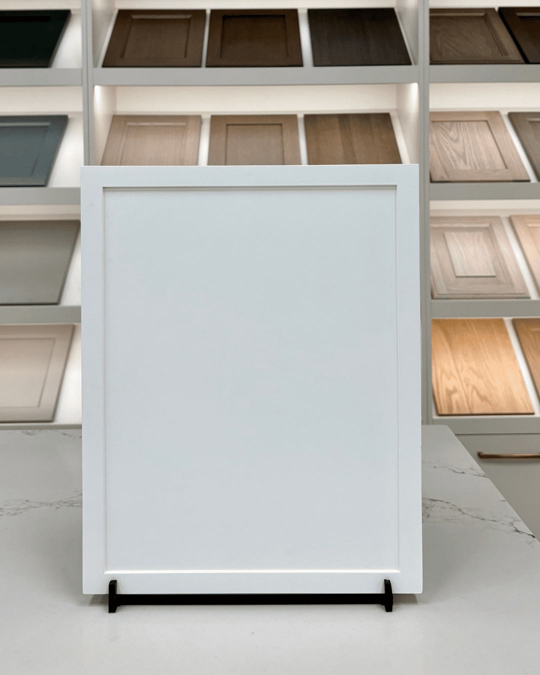 A modern white Slim Shaker cabinet door with thin, minimalist stiles and rails surrounding a recessed center panel, shown on a showroom display stand.