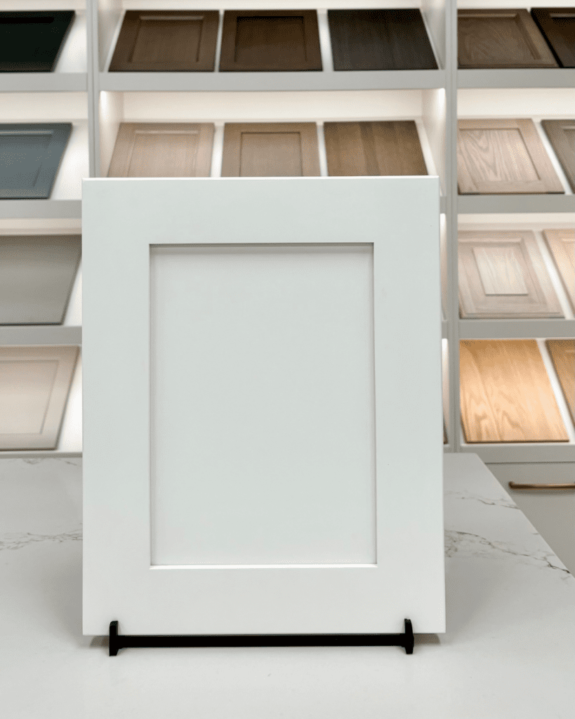 A clean, white Traditional Shaker cabinet door featuring a recessed center panel and wide stiles and rails, displayed on a countertop in a kitchen showroom.