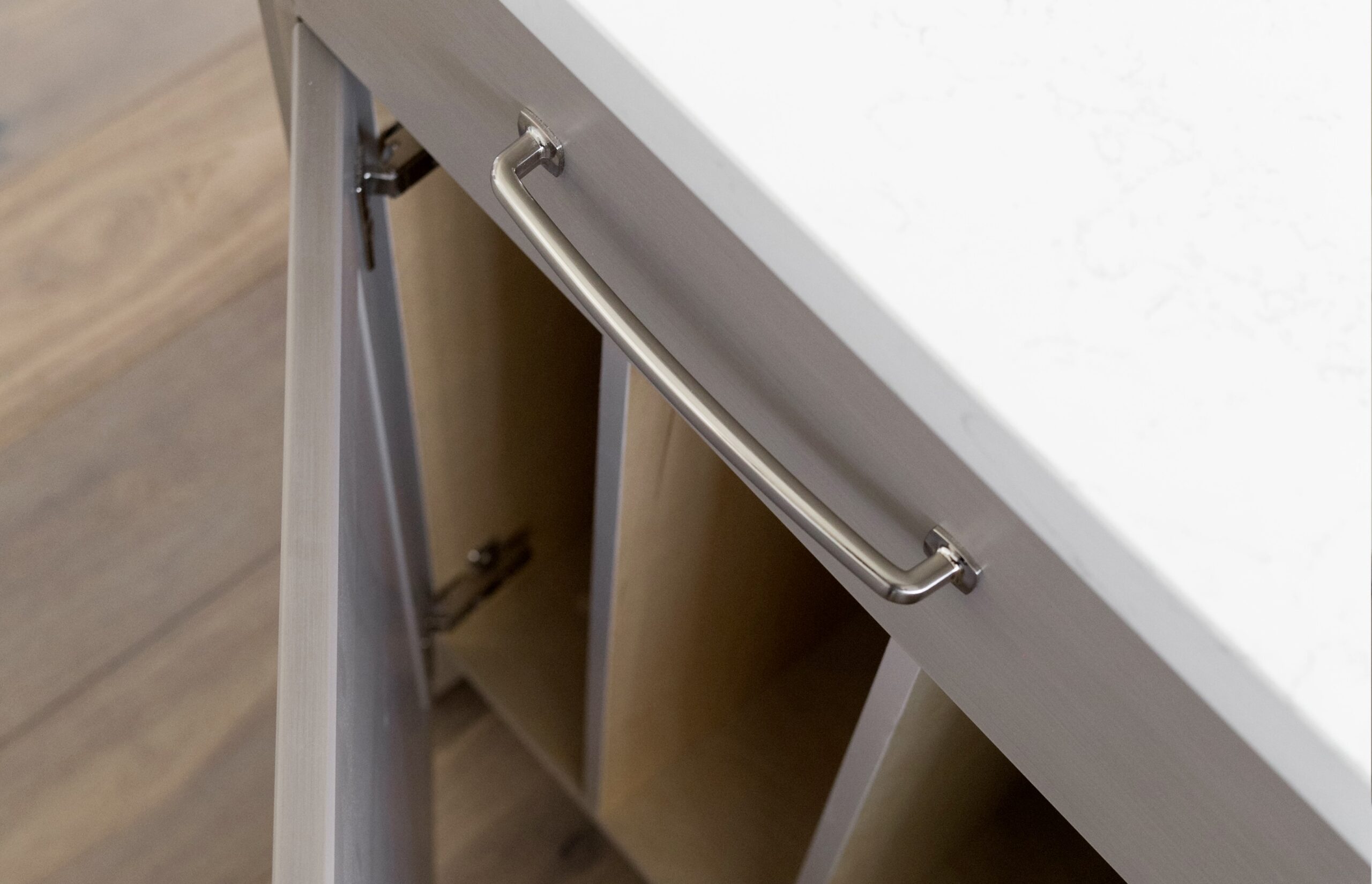 High-angle detail shot of a modern grey cabinet door slightly ajar with premium soft-close hinges, a sleek brushed metal handle, and a white quartz countertop.