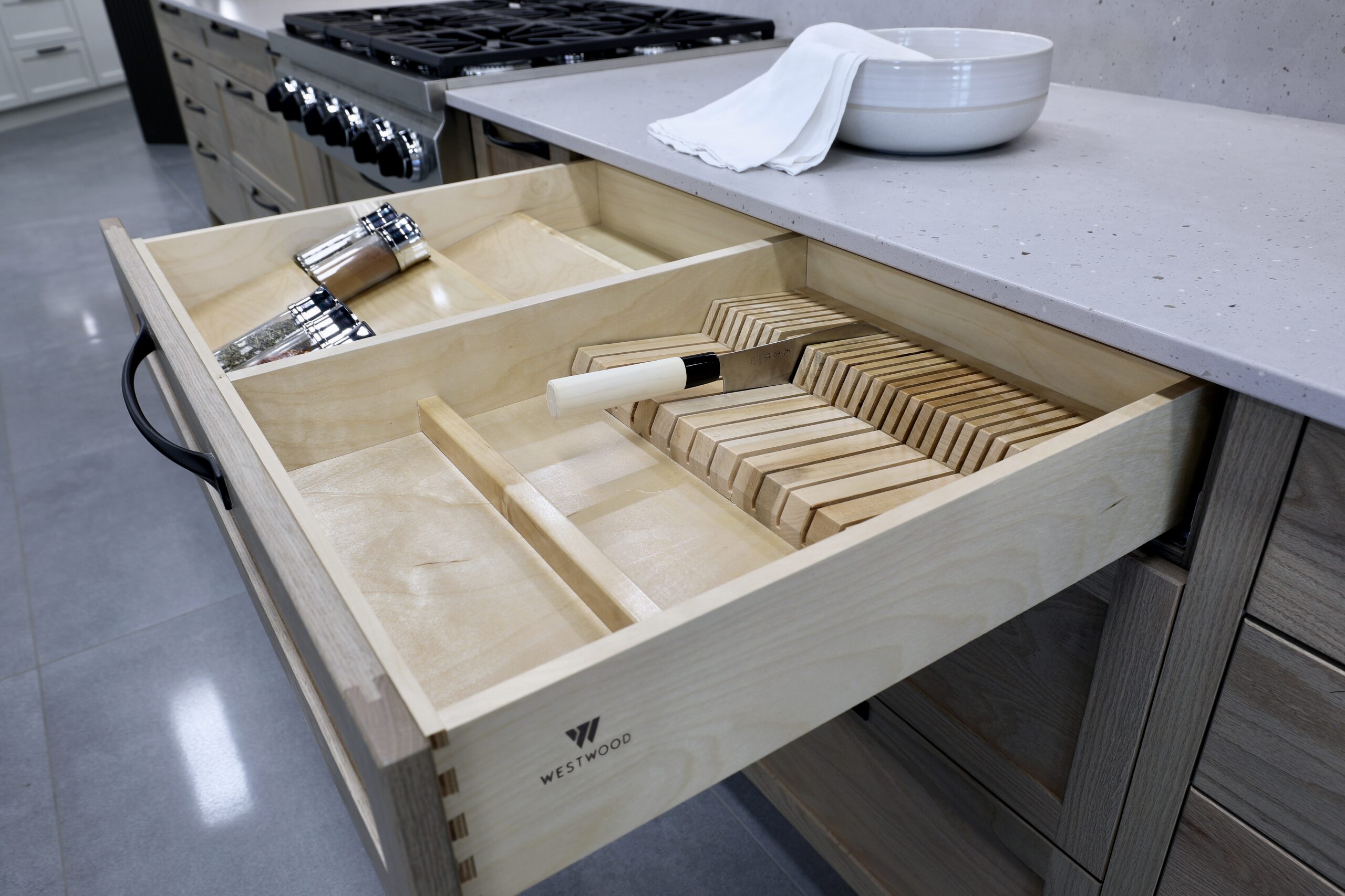 An open custom light wood kitchen drawer from Westwood Cabinetry featuring a built-in knife block with a chef's knife and a dedicated spice rack organizer near a professional range.