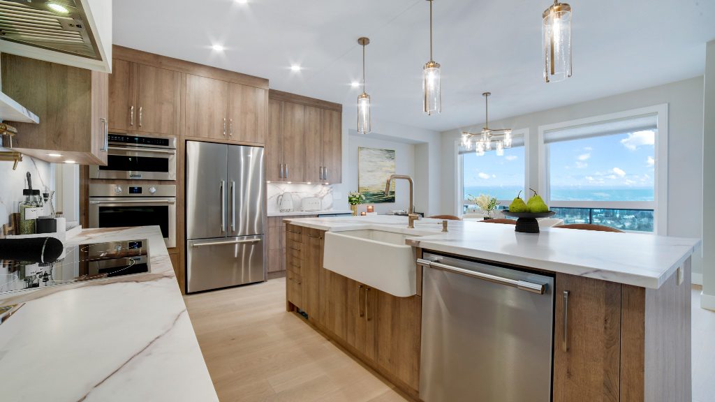A bright, modern kitchen by Westwood designer Anne Woods featuring warm wood custom cabinets with a vertical grain. A large white quartz island sits in the center with a white apron-front sink and gold hardware, set against a backdrop of integrated stainless steel appliances and a large window overlooking a scenic landscape.