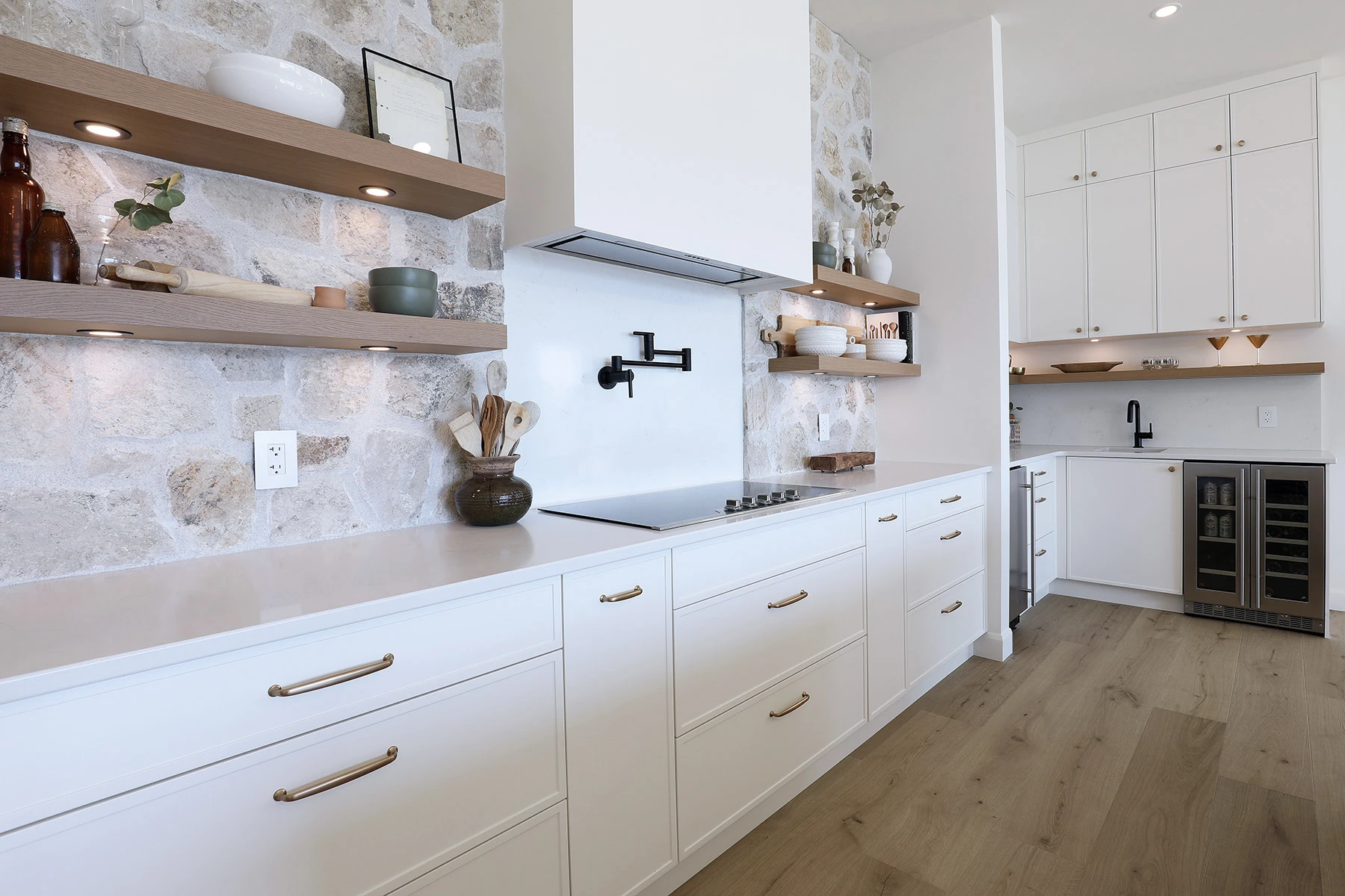 from the Westwood Custom cabinet series collection. A bright, modern rustic kitchen featuring a focal wall made of light-coloured natural stone. Two thick, floating white oak shelves with integrated under-cabinet puck lights are mounted on the stone wall, holding various dishes and decor. A sleek, white minimalist range hood sits above an induction cooktop with a black wall-mounted pot filler. Below, long white drawers with thin brass handles stretch across the counter, transitioning into a secondary wet bar area with a small sink and a dual-zone beverage fridge in the background.