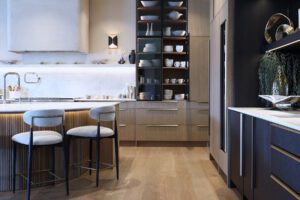 A luxurious contemporary kitchen featuring vertical-grained light oak cabinetry mixed with dark charcoal lower cabinets. The main wall includes a large, smooth plaster range hood over a slab backsplash, flanked by a tall glass-front display cabinet showcasing organized white dishware with integrated lighting. In the foreground, a curved kitchen island with vertical wood integration is paired with two modern barstools featuring black legs and curved white upholstered back