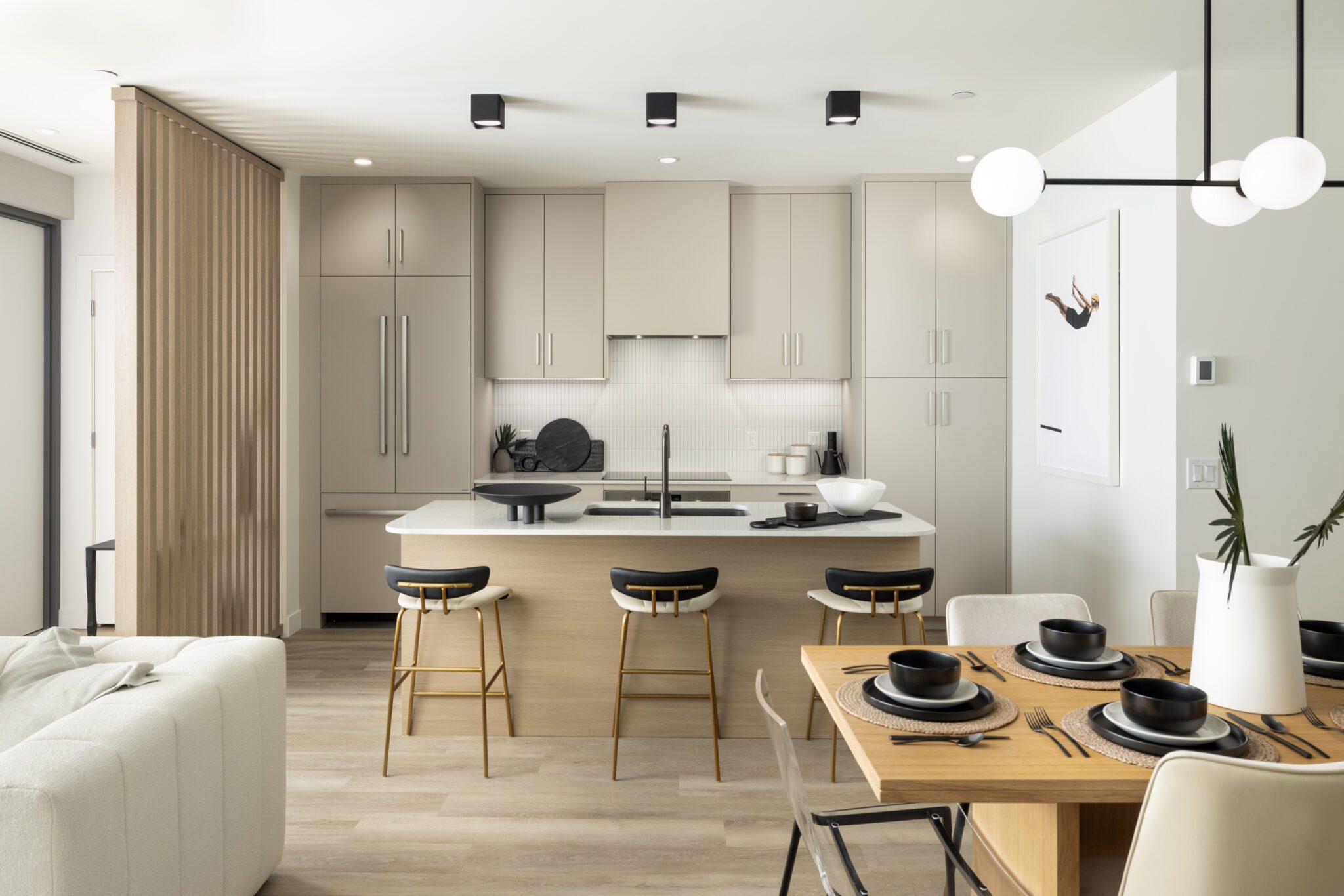 A bright, open-concept kitchen and dining area with a minimalist aesthetic. The kitchen features flat-panel cabinets, a matching integrated refrigerator, and a central island with a white countertop and light wood base. Three modern barstools with black seats, white backs, and gold legs line the island. In the foreground, a light wood dining table is set with black bowls on round woven placemats under a modern black and white globe pendant light. A vertical wood slat room divider stands to the left.