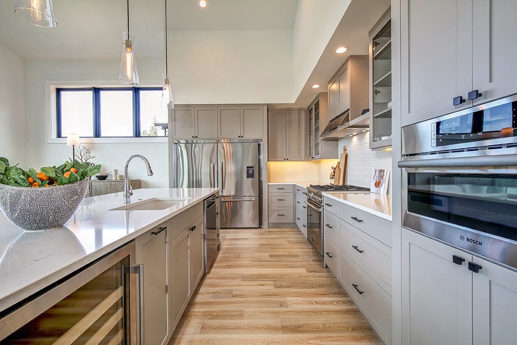 A spacious, high-ceiling kitchen featuring light grey Shaker-style cabinetry and white quartz countertops with subtle veining. A long central island with a built-in sink and a stainless steel wine cooler runs down the left side, illuminated by three hanging glass pendant lights. The back wall features a large stainless steel double-door refrigerator, a gas range with a stainless steel hood, and a wall unit on the right housing a built-in Bosch oven. The floor is made of light-coloured hardwood planks.