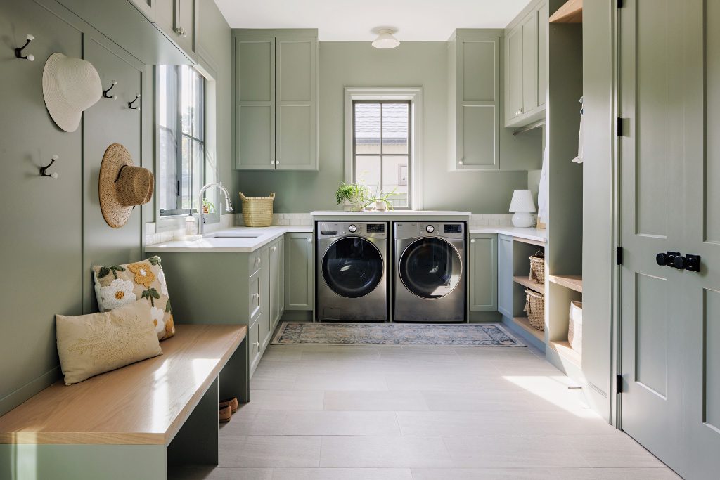 Custom sage green laundry room featuring built-in cabinetry, an integrated mudroom bench with coat hooks, and high-efficiency appliances under a continuous folding counter.