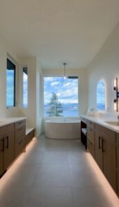 Modern ensuite featuring custom bathroom cabinetry in Kelowna with integrated under-cabinet lighting, dual vanities, and a freestanding tub framing a scenic outdoor view.