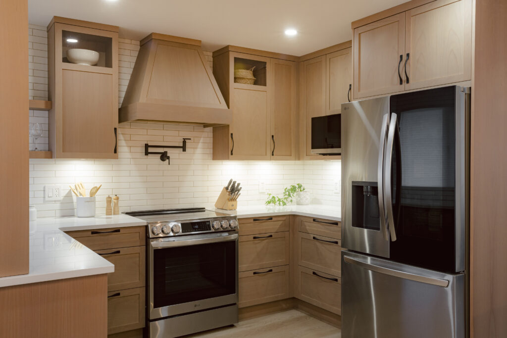 A bright, compact kitchen featuring light wood custom cabinetry, white quartz countertops, a matching range hood, and stainless steel appliances.
